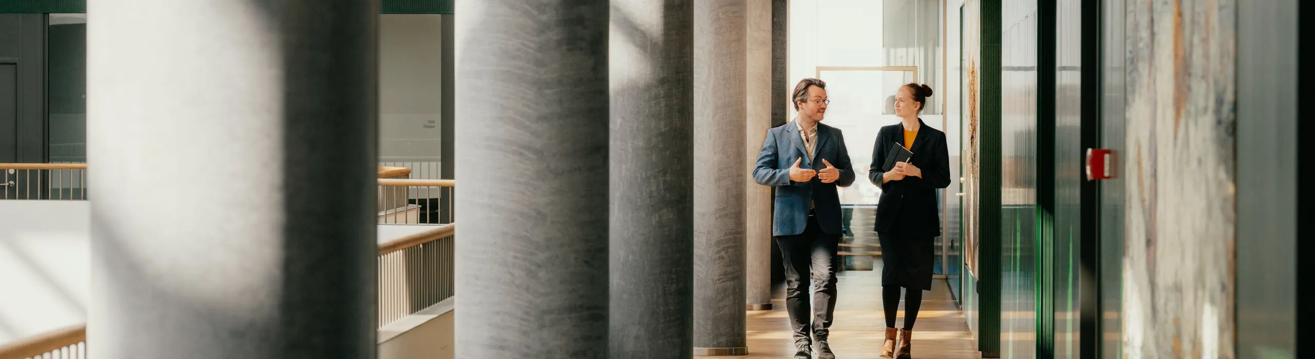 Two people walking and talking in a bright hallway with white acoustic modular ceilings, large columns and artwork, illuminated by sunlight streaming through windows.