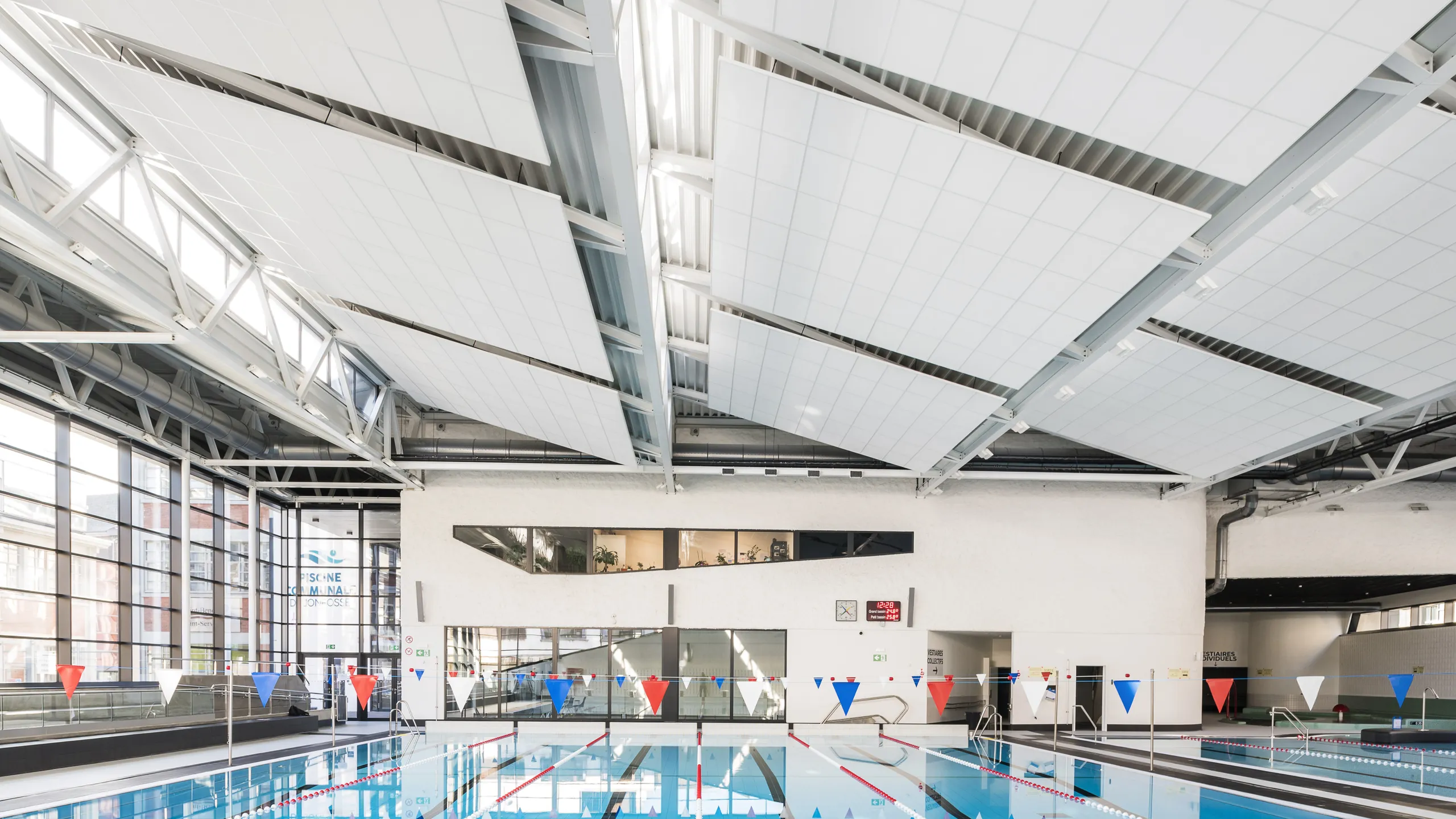Indoor swimming pool with suspended white acoustic ceiling rafts mounted between exposed structural beams, above a lane pool with flags, large windows, and a bright, open pool hall.
