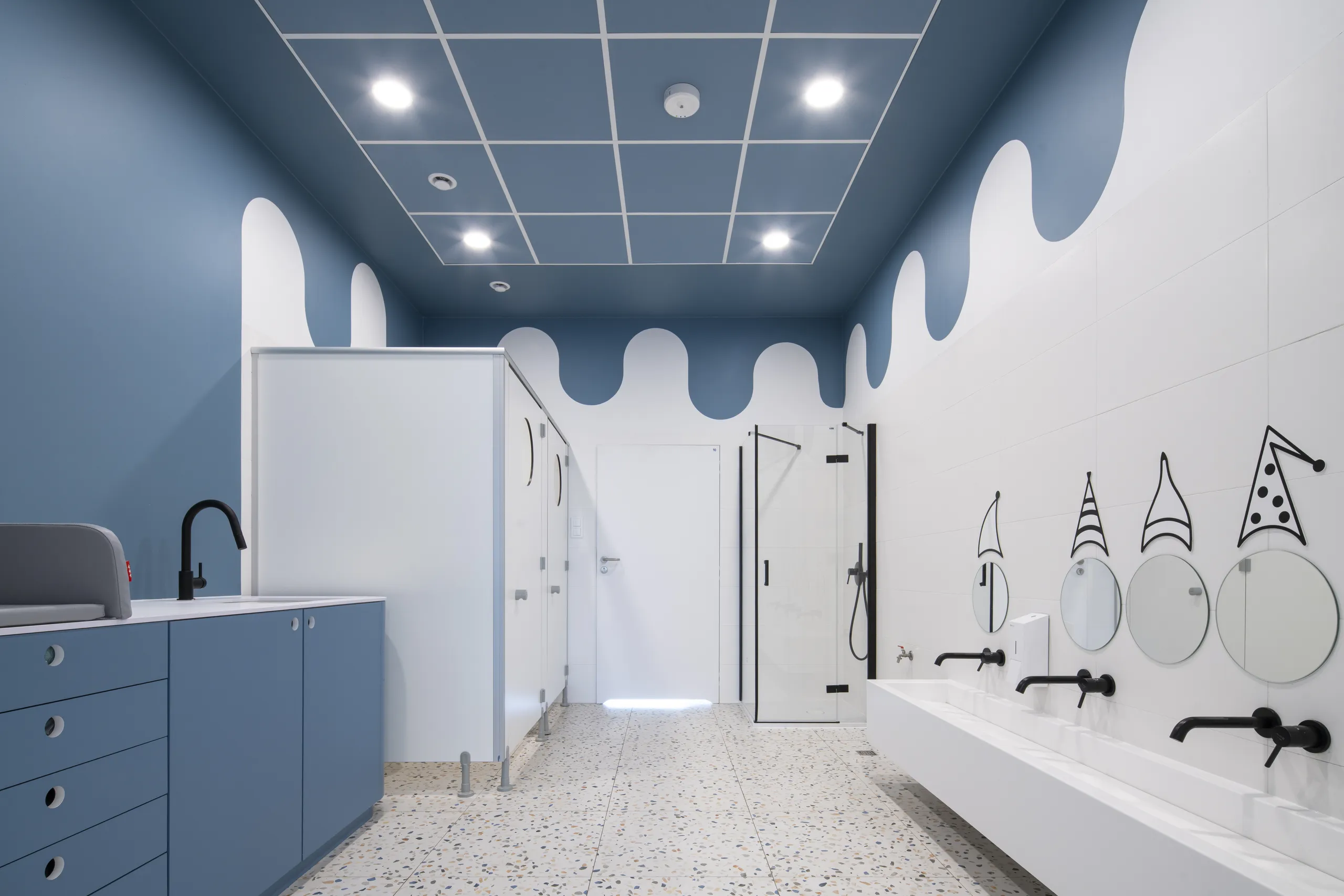 A bright children’s bathroom featuring a blue acoustic ceiling tile grid with recessed lights, wave-pattern wall graphics, circular illustrated mirrors, and child-sized basins and cubicles.
