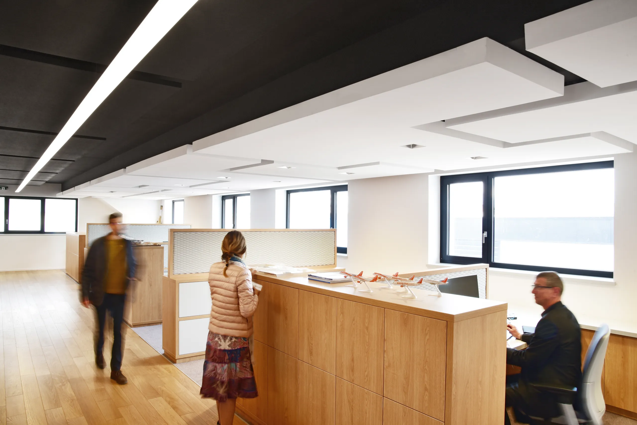 Bright office with large floating acoustic rafts suspended under a dark ceiling, above wooden desks and high windows