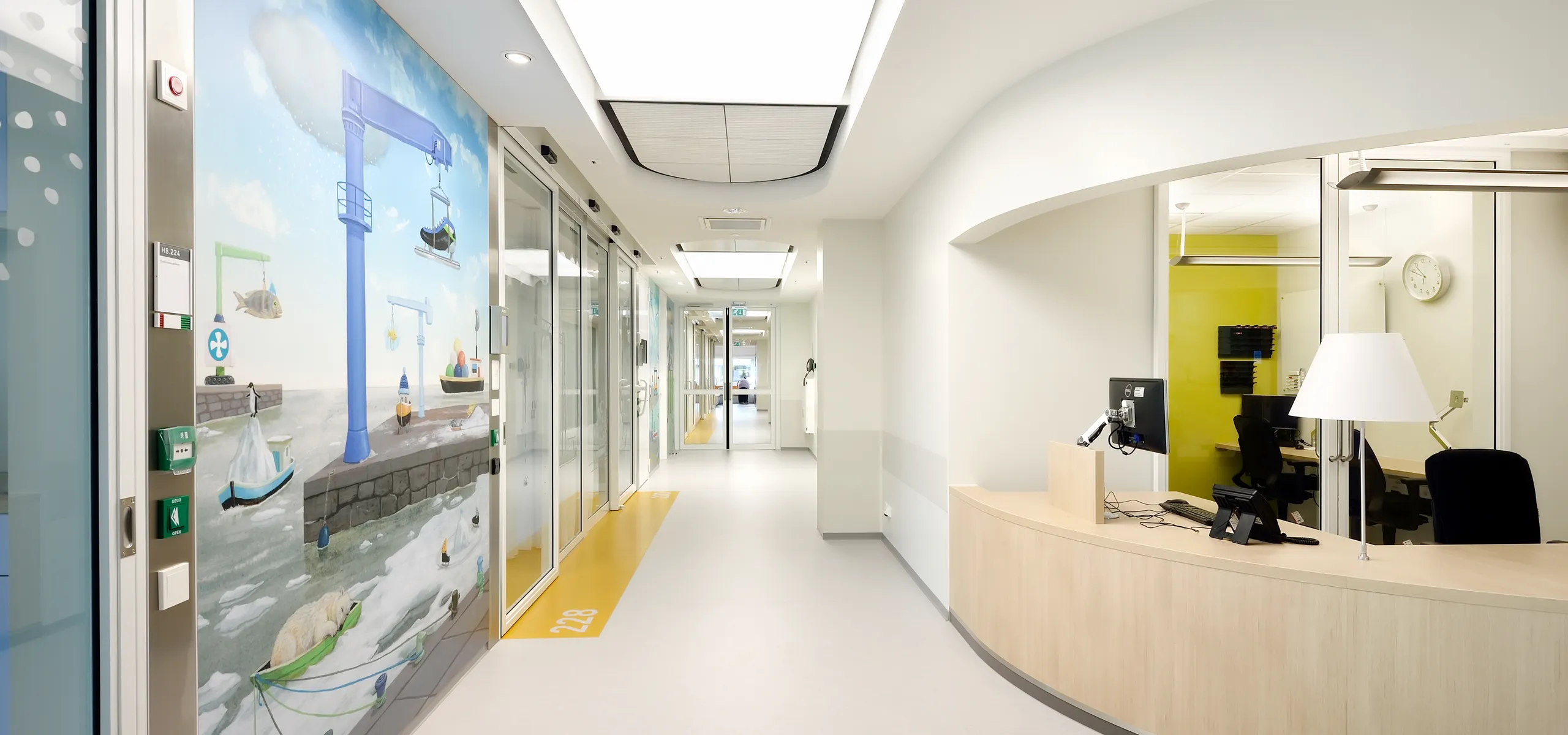Hospital corridor featuring a nurse station with wall art, a curved reception desk and a large illuminated acoustic ceiling panel running down the hallway.