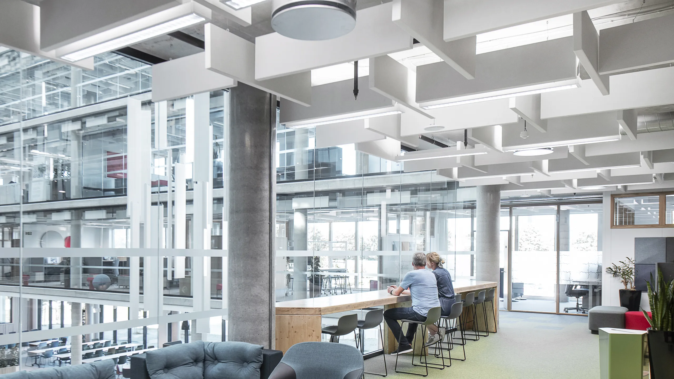 Modern office lounge with a baffle office ceiling, sofas, chairs, and bar seating. Two people are sitting at the bar. Glass walls overlook an open interior space.