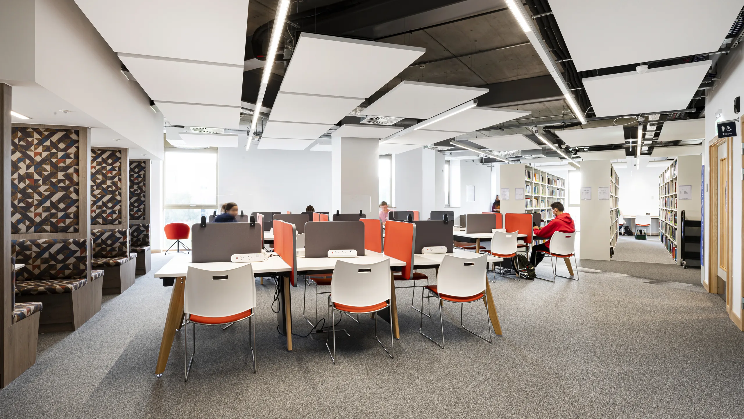 Library study area with suspended white acoustic ceiling panels, desks with study stations, and shelving units arranged in an open, modern learning environment.