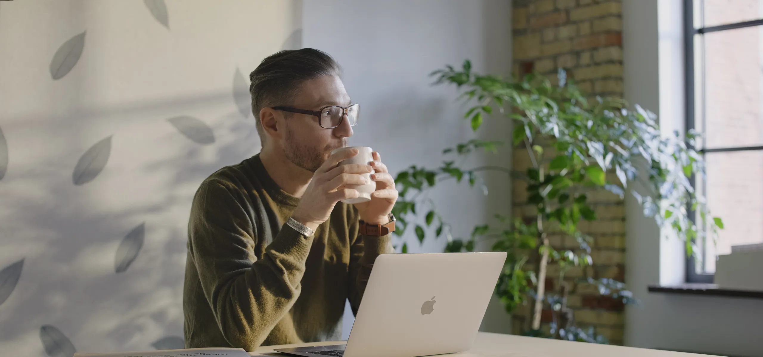"Man drinking coffee at his desk with a laptop, seated in front of an acoustic wall panel featuring a natural leaf pattern.