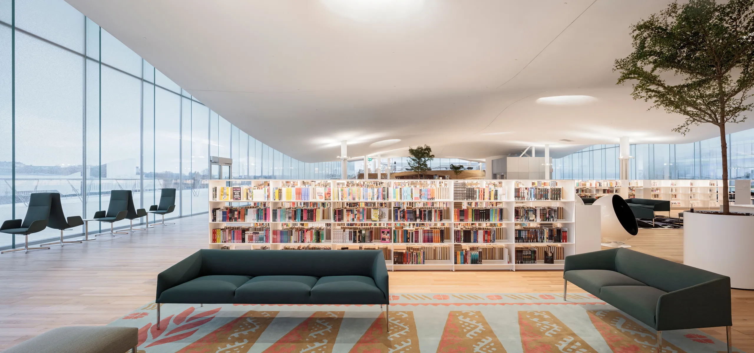 A bright public library with a seamless white acoustic ceiling, large glazed façade, and wide timber steps filled with book.