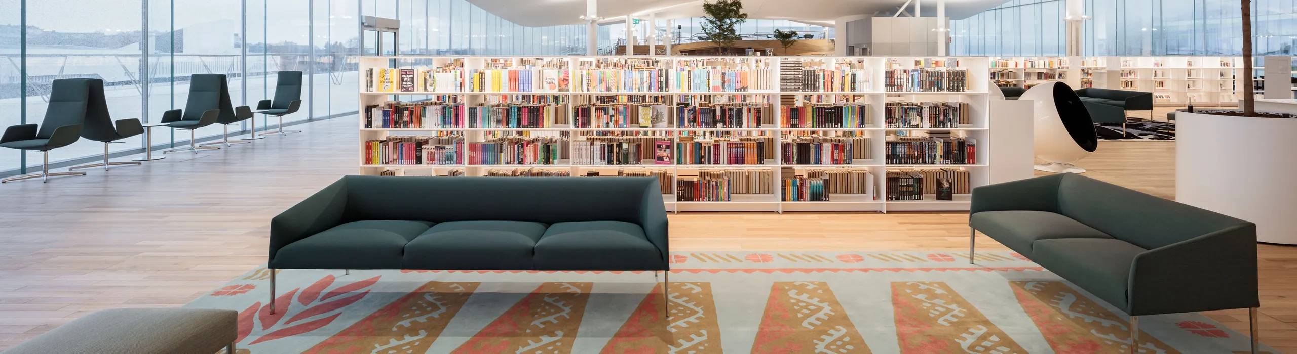 A bright public library with a seamless white acoustic ceiling, large glazed façade, and wide timber steps filled with book.