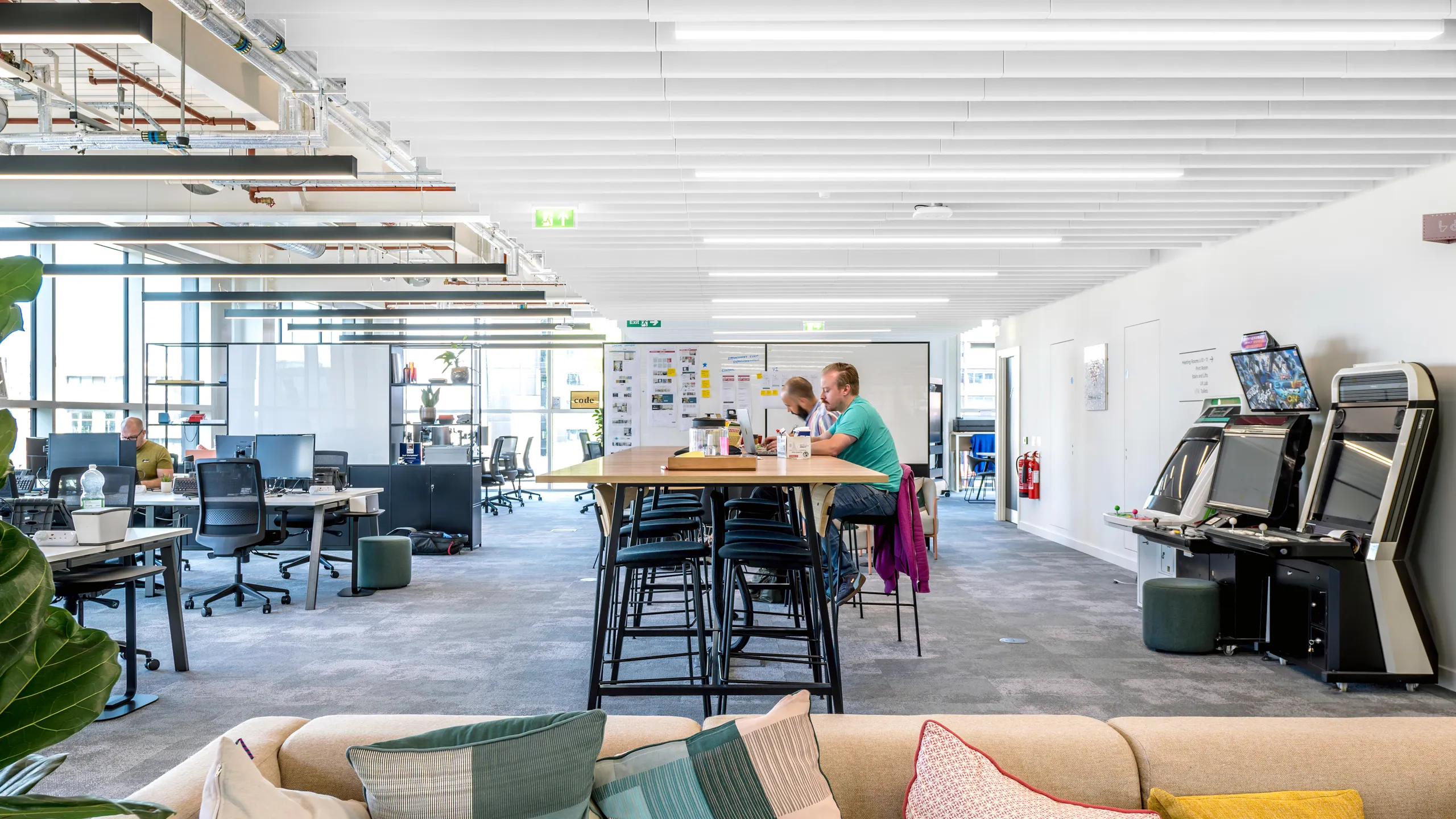Open-plan office workspace with desks, collaboration tables, and white acoustic baffle ceiling panels above the interior.