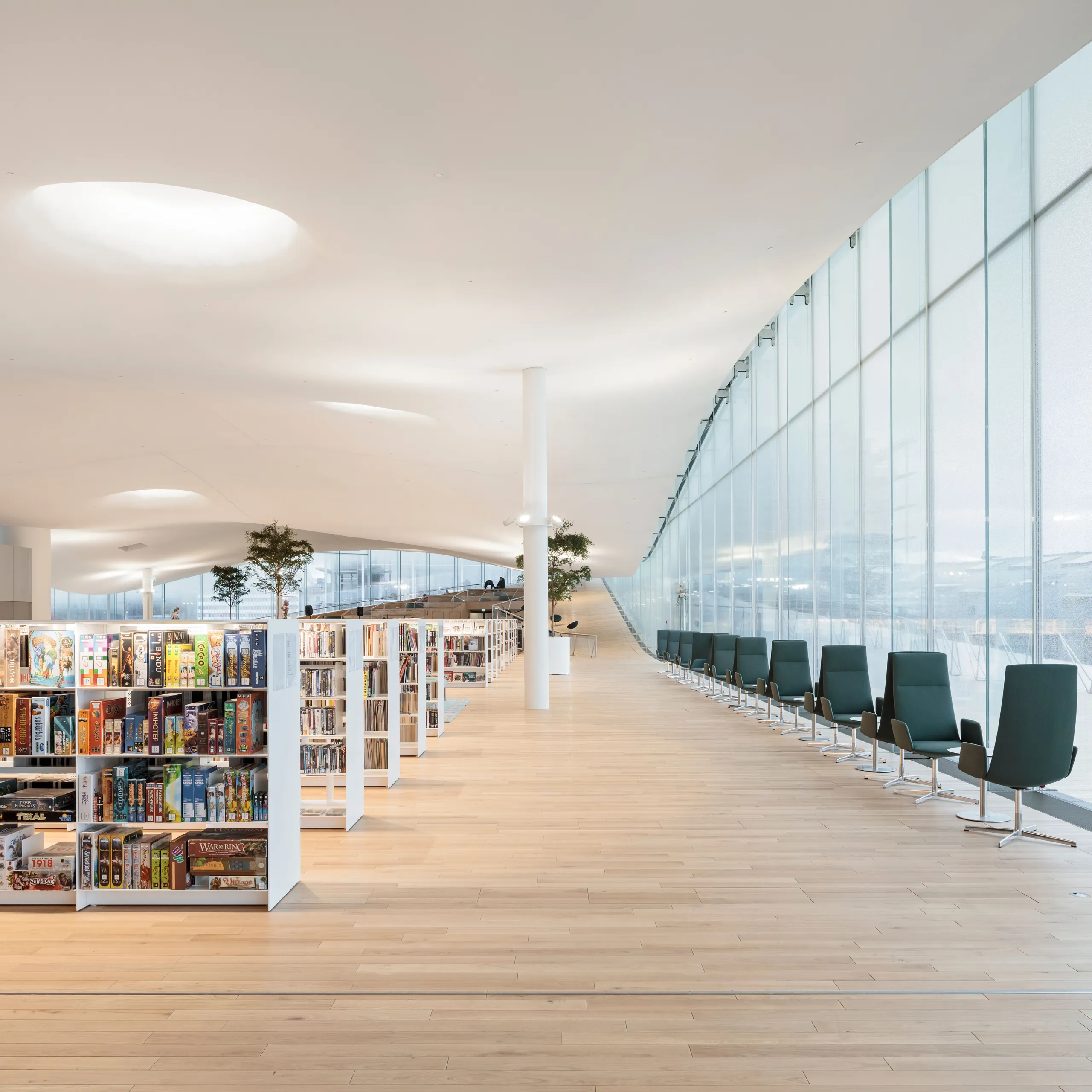 Library interior with low bookshelves, seating along a full-height glass façade, indoor trees, and a smooth white acoustic ceiling with circular skylights.