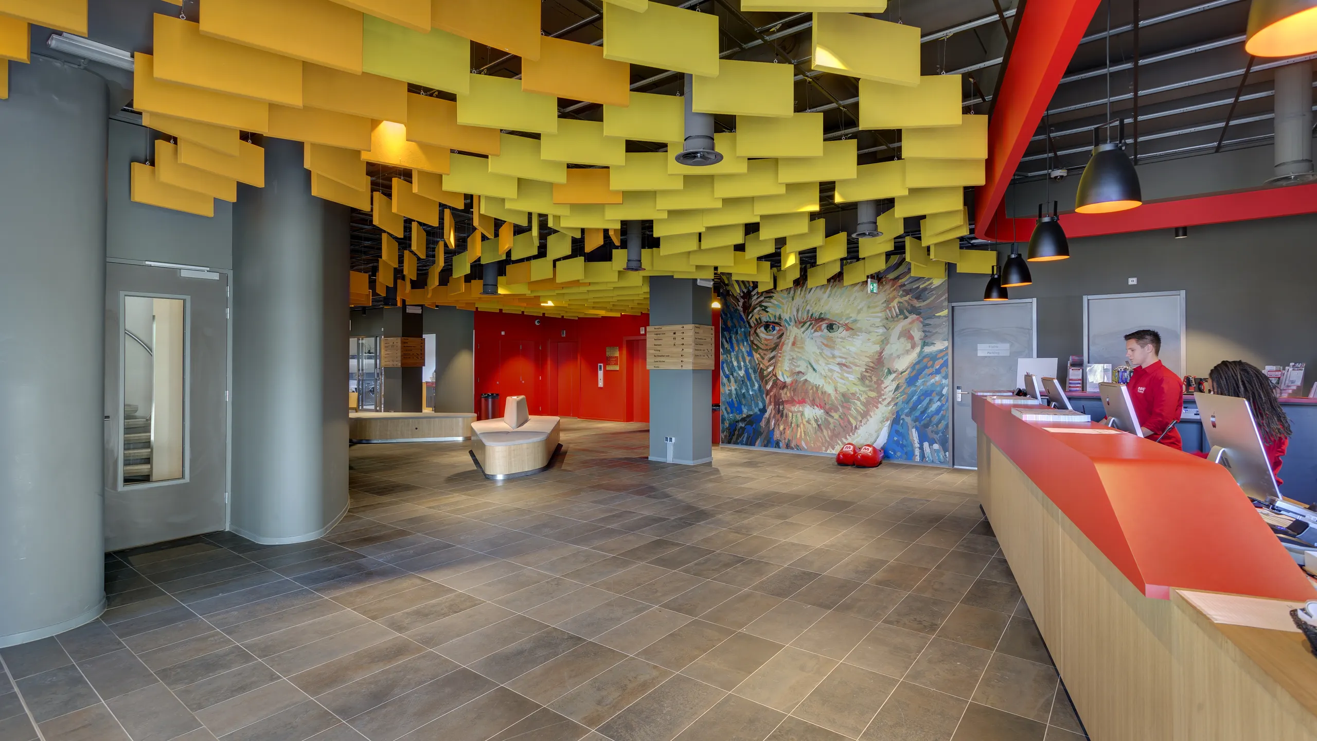 Reception area with suspended yellow acoustic baffles forming a dynamic ceiling, red reception desk, and large mural artwork on the wall behind the lobby space.