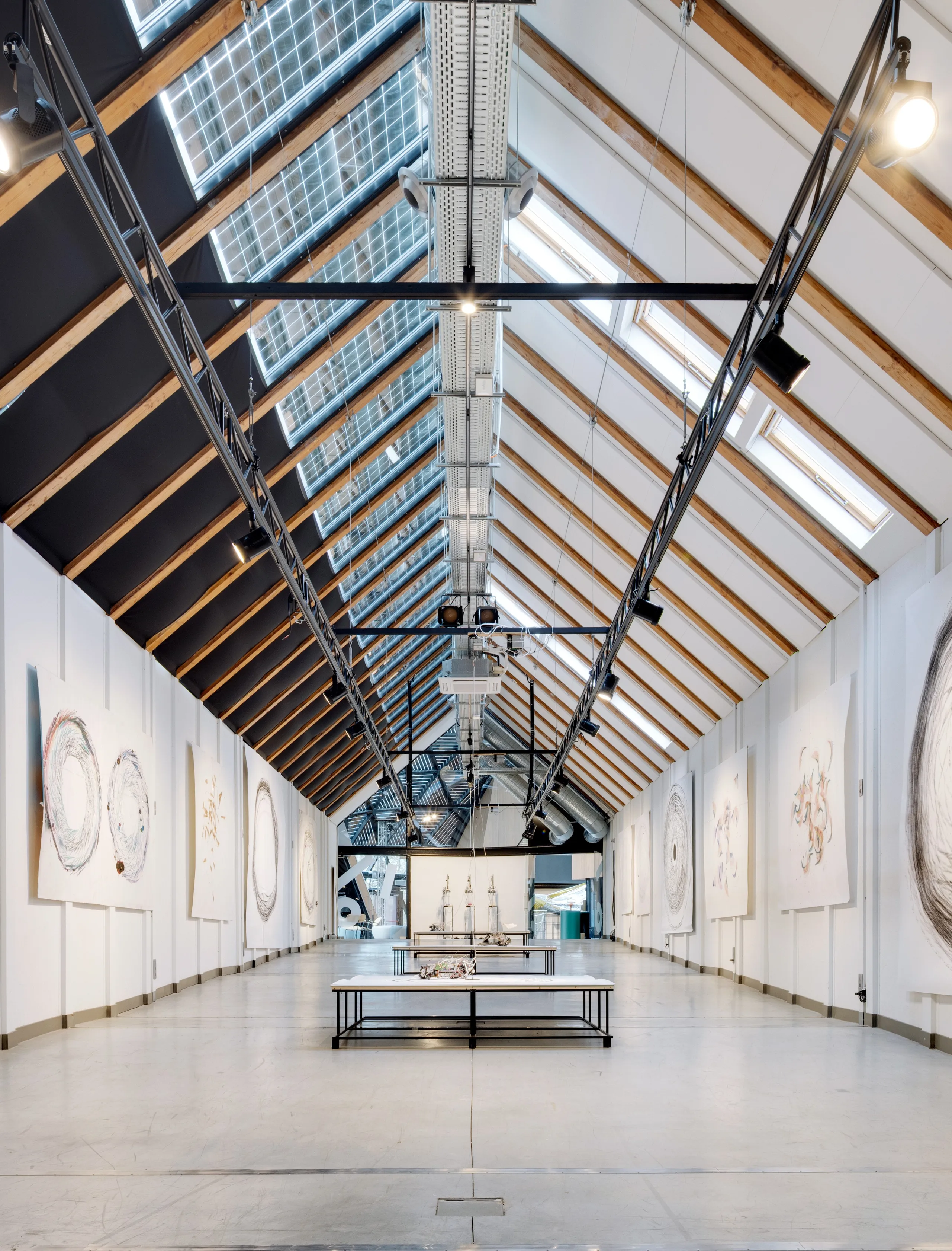 Art gallery interior with a high pitched roof, timber beams, large skylights, suspended lighting tracks, and artworks displayed along white walls around a central exhibition space.