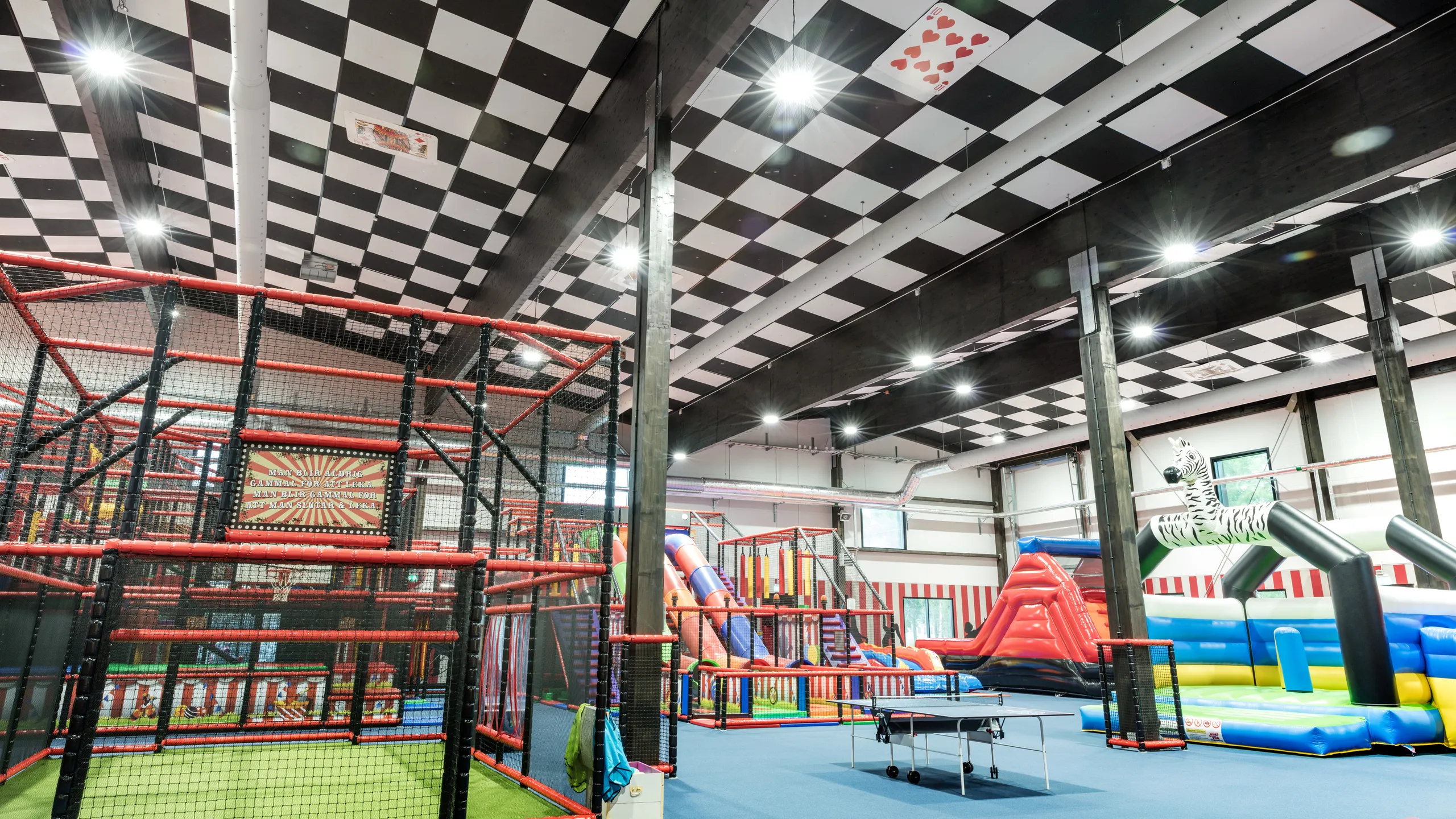 Indoor play centre with a black and white checkerboard acoustic ceiling, climbing frames, slides, trampolines, and inflatable play structures.