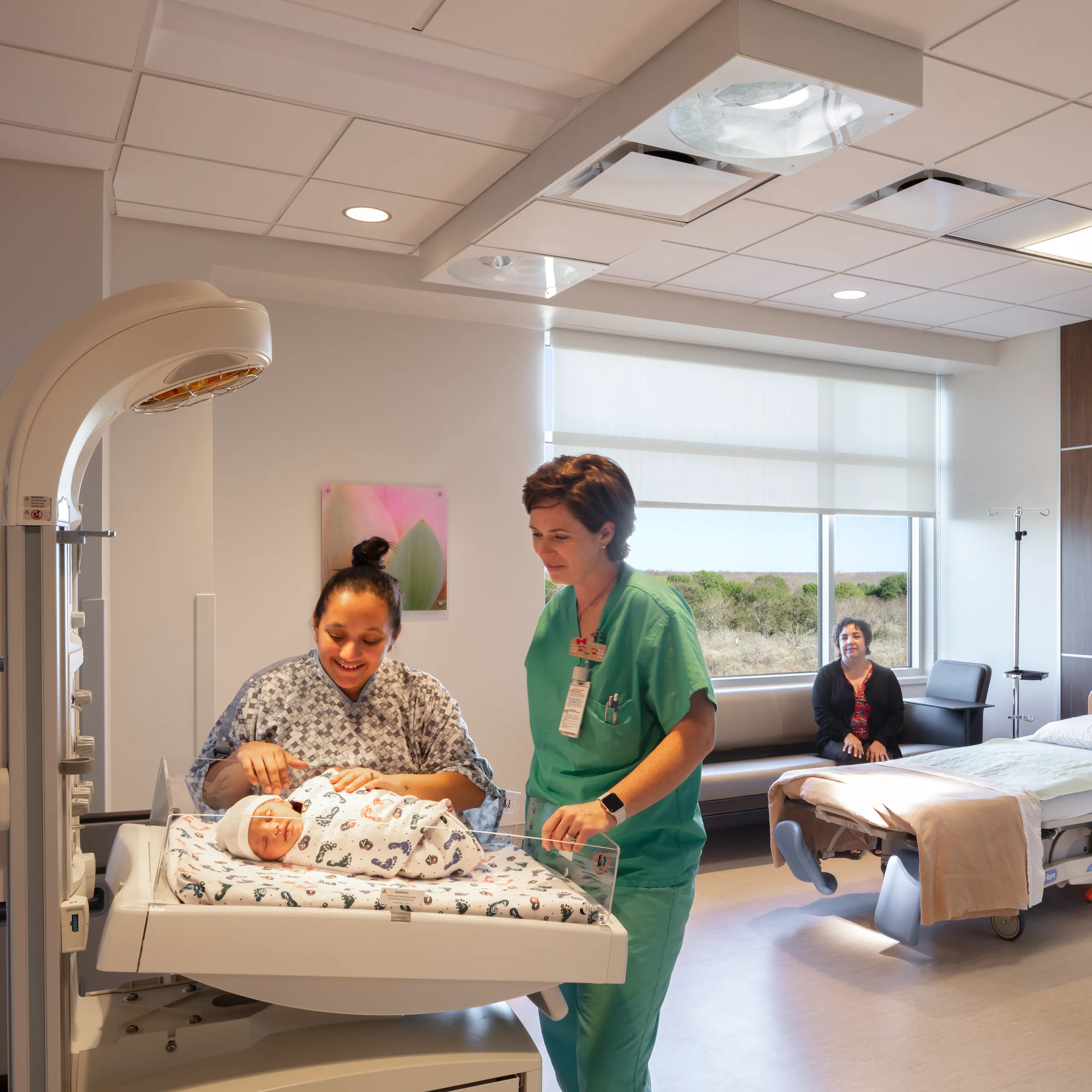 A maternity patient room with a new mother, nurse and newborn under a warming station, featuring a suspended acoustic ceiling grid and soft natural daylight.