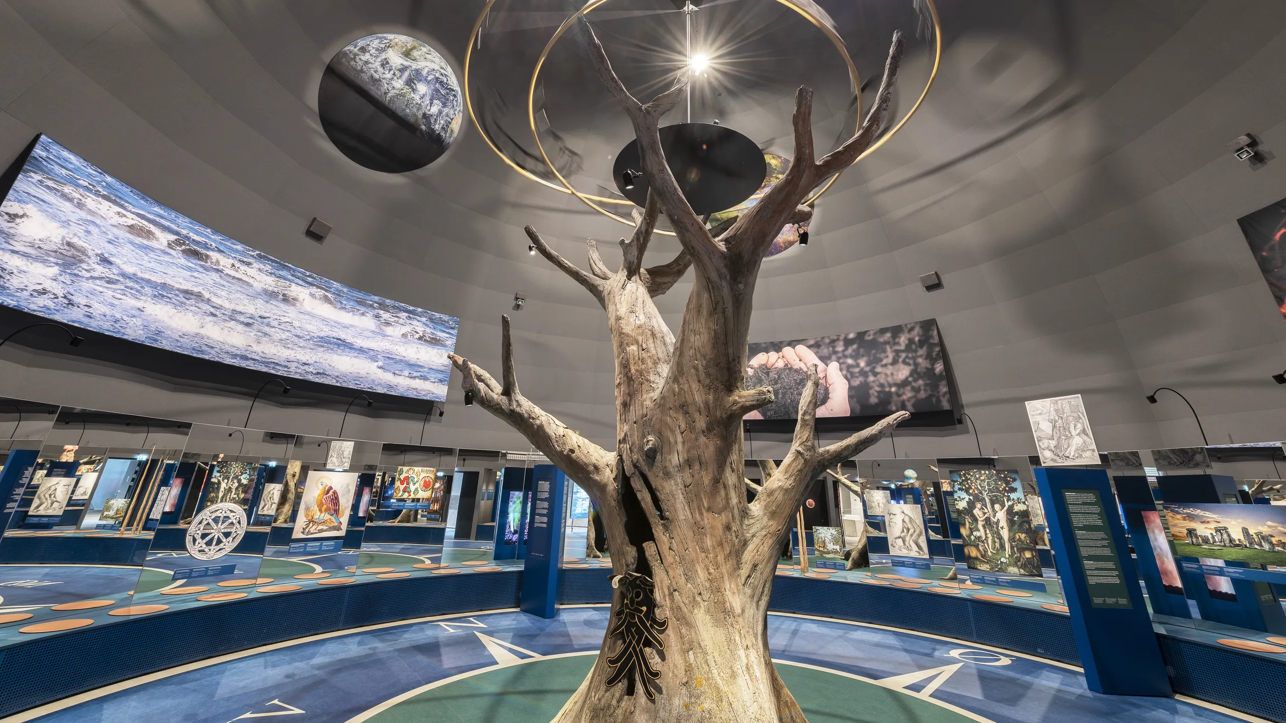 Exhibition space at Naturkraft in Ringkøbing with a grey acoustic ceiling, central tree installation, suspended planet models, and interactive displays arranged around the circular gallery.