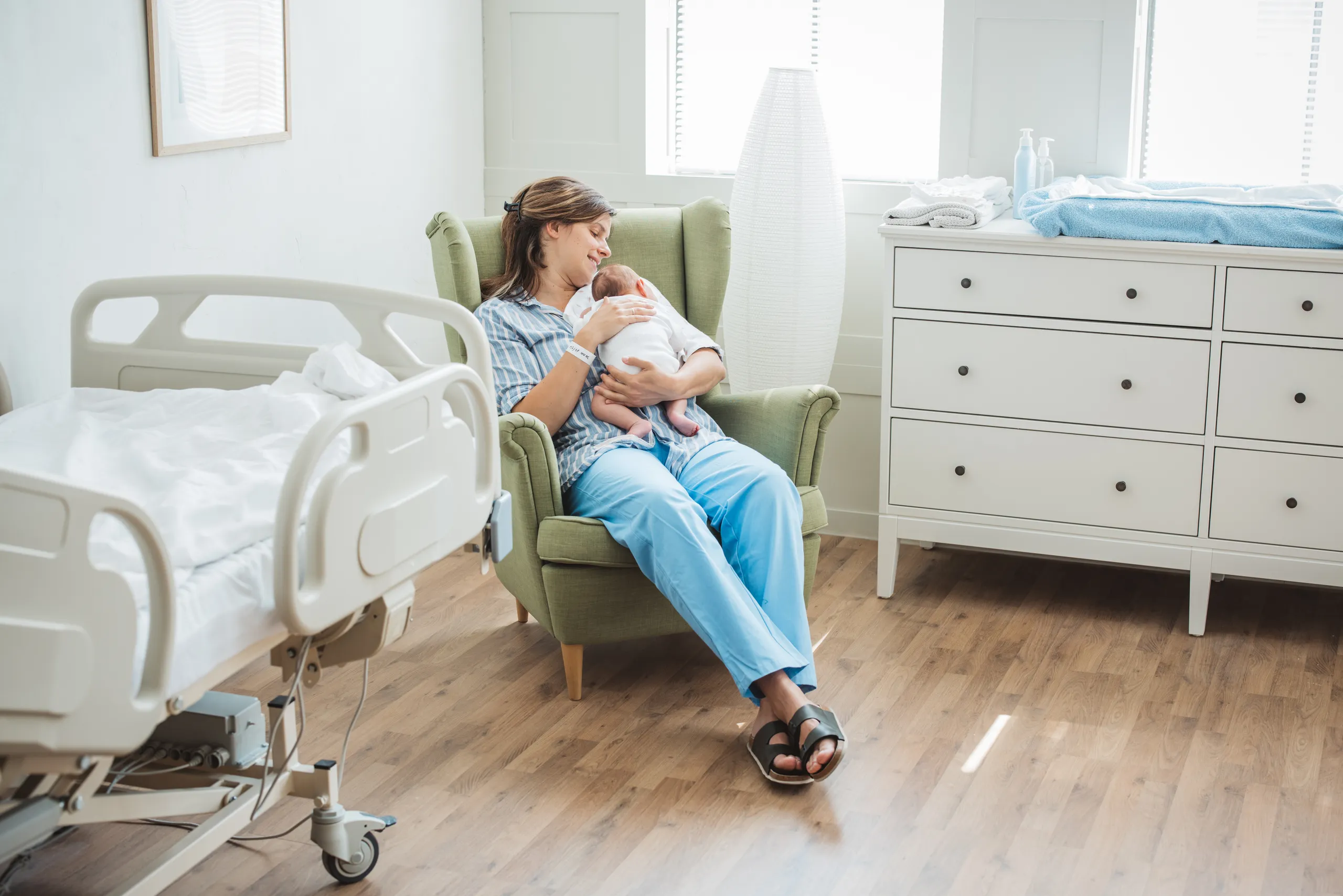 Mother holding newborn in a cozy hospital room with a bed and dresser, sitting in a green armchair by the window.