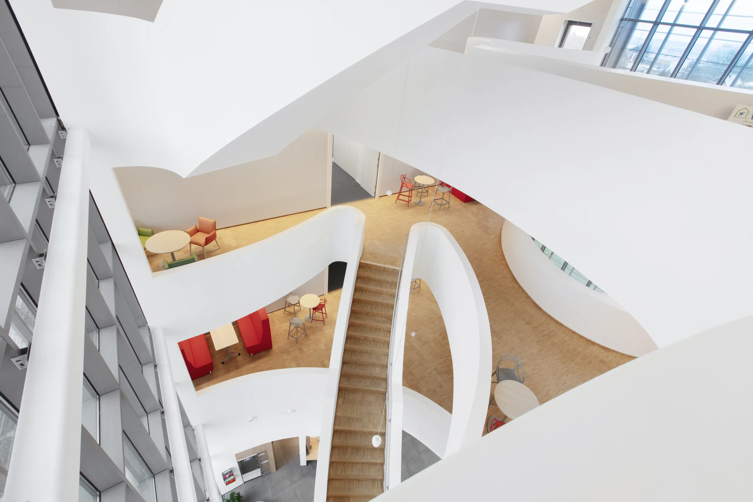 Multi-storey atrium with a monolithic acoustic ceiling, curved white walkways, wooden floors, and small seating areas viewed from above