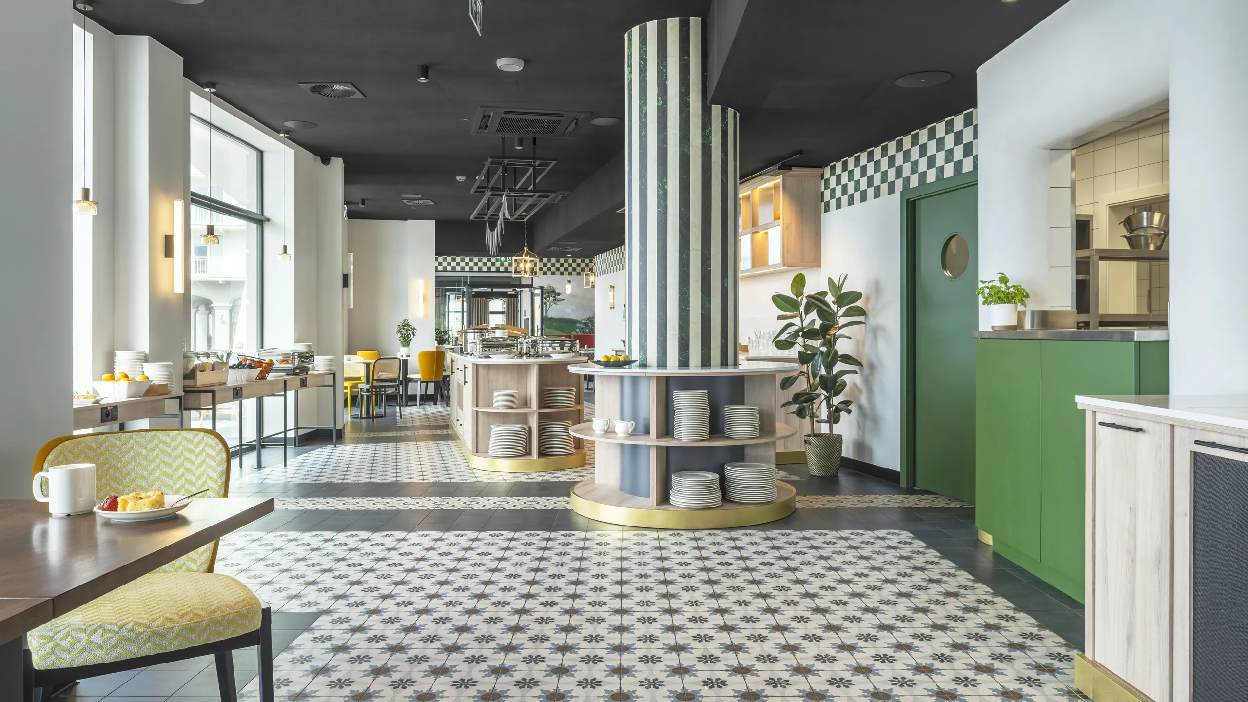 Bright, modern café interior with patterned floor tiles, black suspended ceiling, green and yellow chairs, wall art, hanging lights, and plants for decoration.